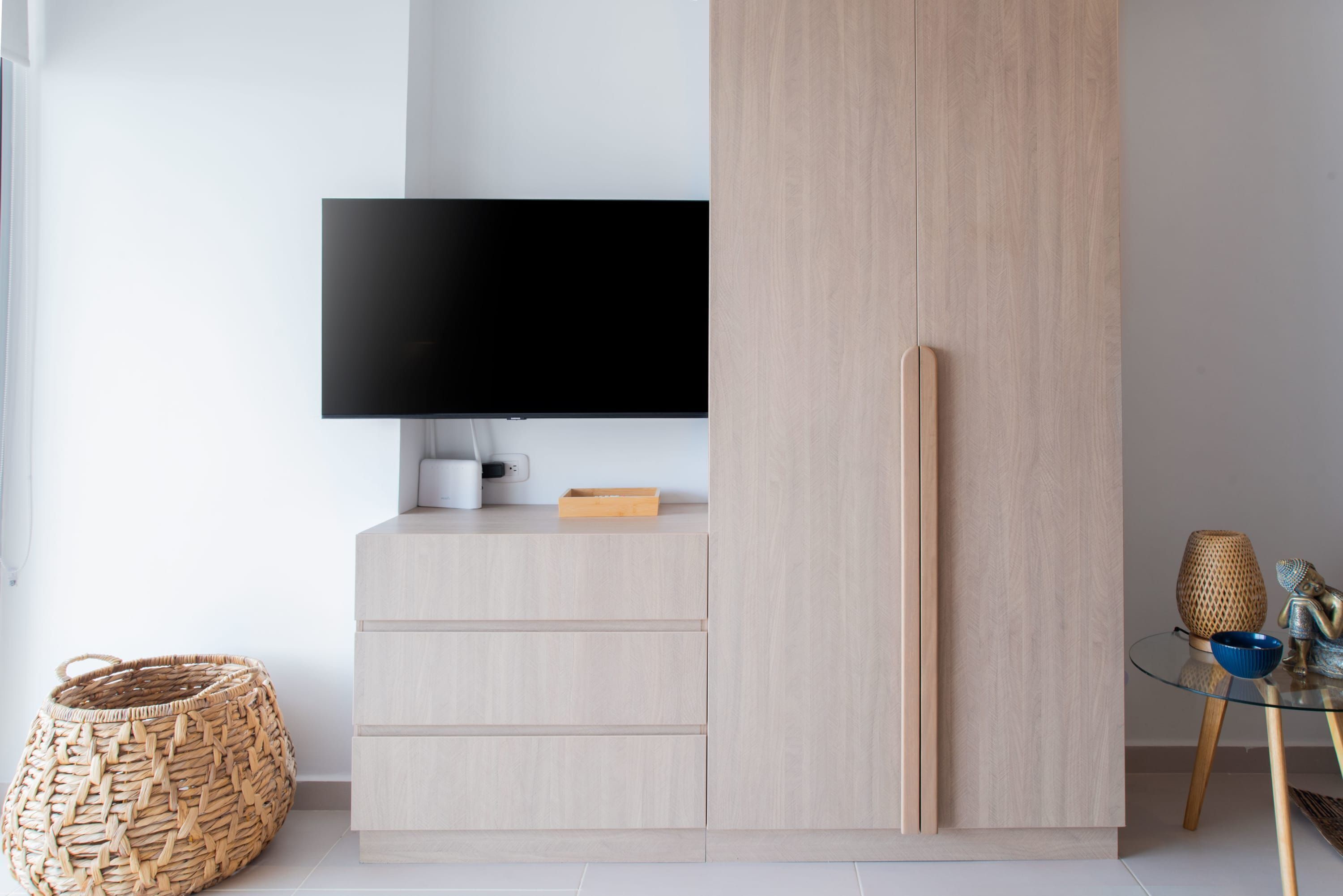 A modern room with a flat-screen TV mounted on a wall, a wooden wardrobe, a small table with decorative items, and a woven basket on the floor.