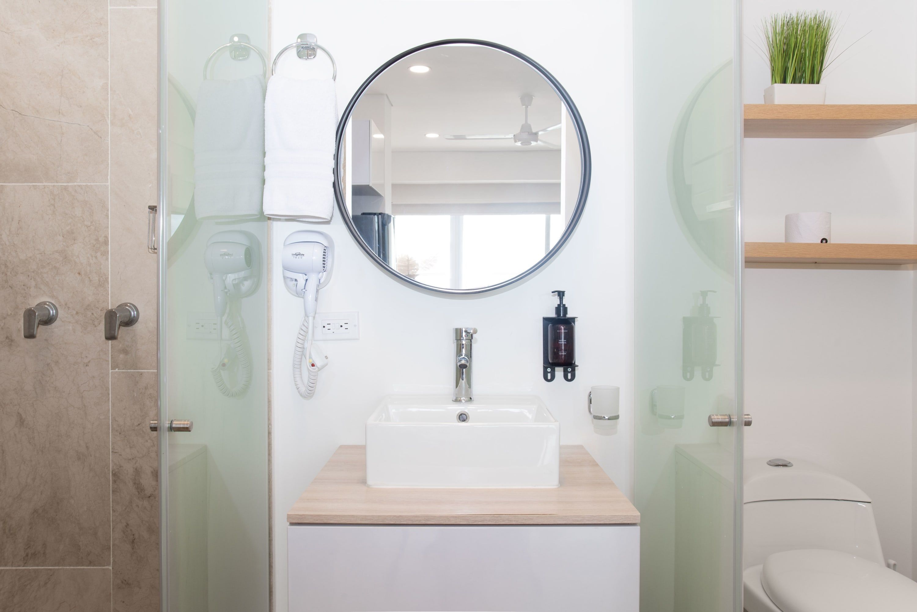 Une salle de bain avec une douche à parois de verre, un lavabo blanc doté d’un plan de toilette en bois, un miroir rond et des toilettes.