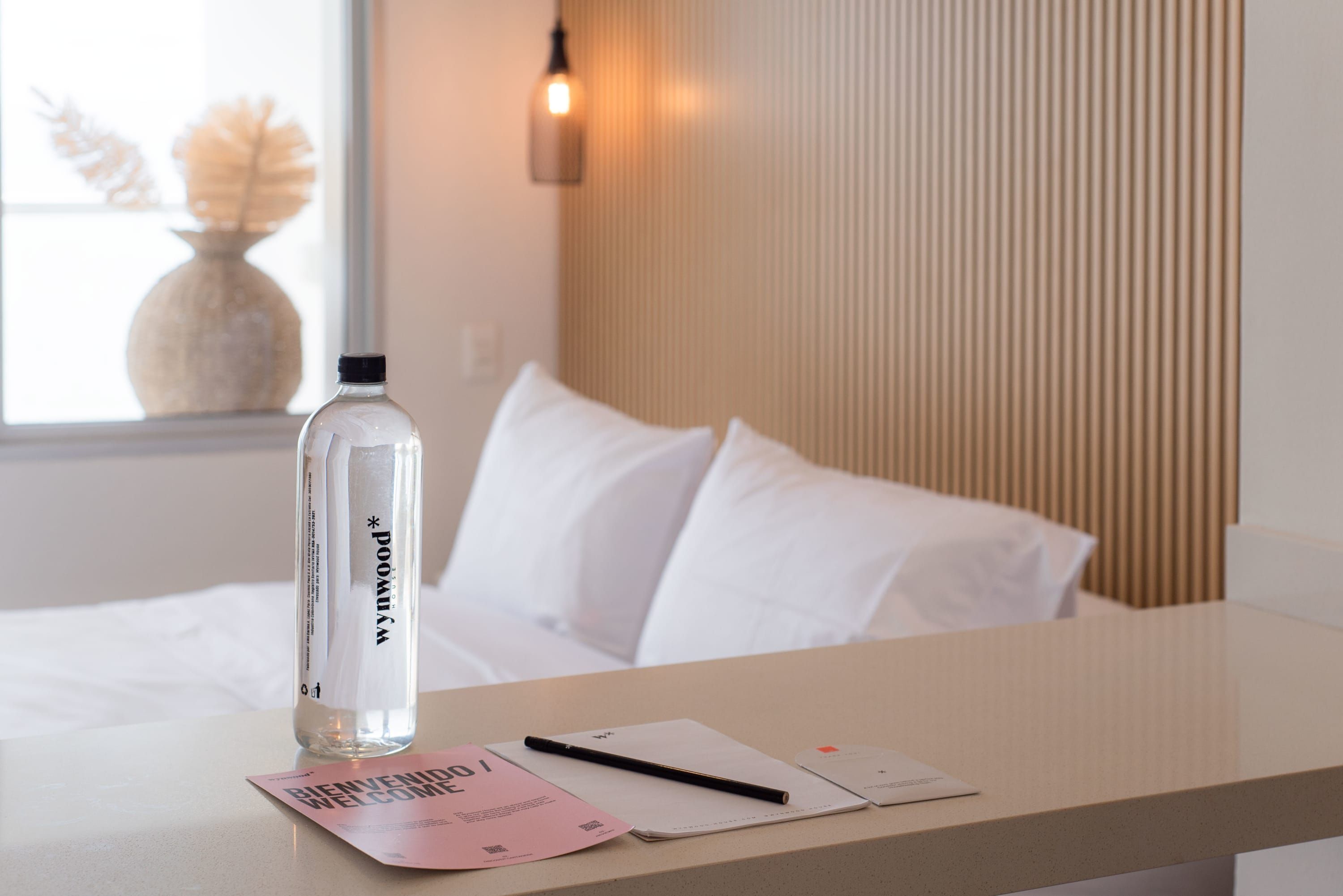 A hotel room with a bed, a bedside table with a water bottle, a notebook, and a welcome card.