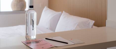 A hotel room with a bed, a bedside table with a water bottle, a notebook, and a welcome card.