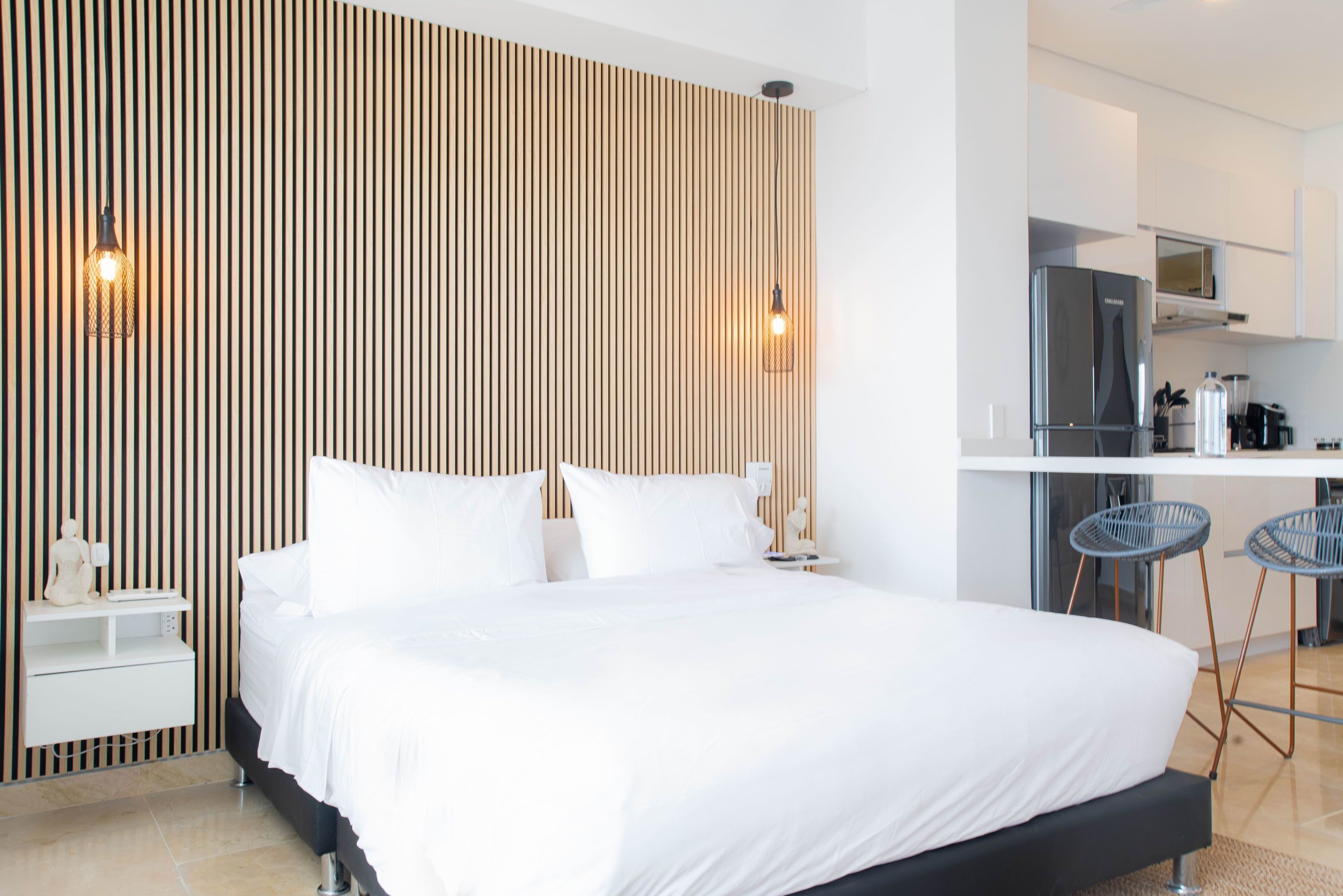 Ett modernt hotellrum med en stor säng, en väggpanel av träribbor, ett litet kök och två barstolar.