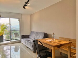 A modern living room with a grey sofa, a wooden dining table, and a black chair. There is a potted plant on the balcony and a bottle on the table. - w Breathtaking 1BR With Terrace at Poblado (Medellín)
