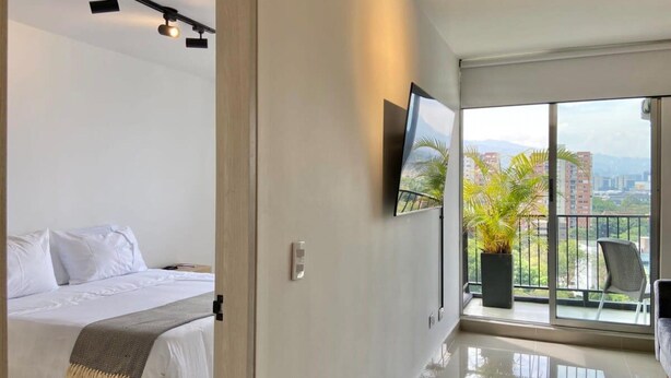 Una habitación de hotel moderna con cama, televisor de pantalla plana, balcón con plantas y vistas a la ciudad.