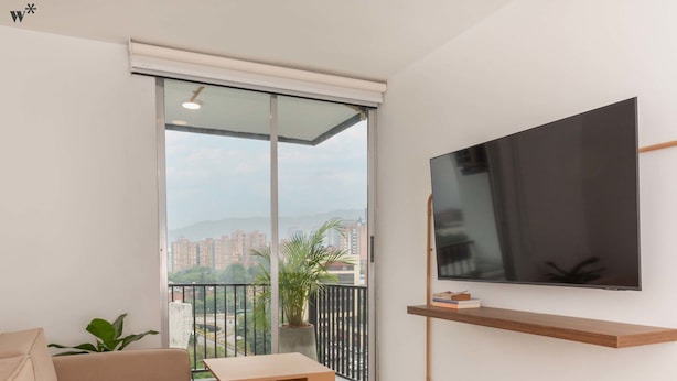 Ein modernes Wohnzimmer mit einer großen Glasschiebetür zum Balkon, einem wandmontierten Flachbildfernseher, einem beigefarbenen Sofa und einem kleinen Holztisch.