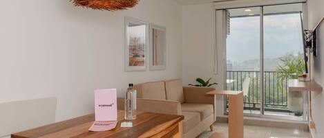 A modern living room with a wooden table, a beige sofa, a wicker pendant light, and a large window with a city view.