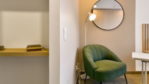 Une chambre moderne avec un fauteuil vert, un miroir rond et une petite étagère en bois où sont posés des livres.