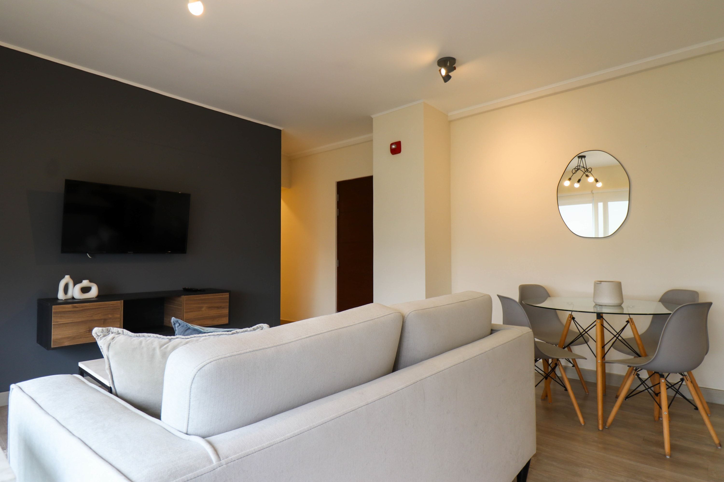 A modern living room with a sofa, a flat-screen TV, a dining table with chairs, and a round mirror on the wall.