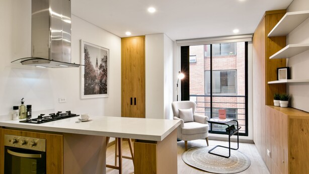 A modern kitchen with a central island, built-in oven, and stainless steel range hood. There is a window with a view of a brick building. - Lovely Loft With Modern Deco in Chapinero (Bogotá)