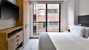 A modern bedroom with a large bed, a TV mounted on the wall, and a view of a brick building through the window. - Lovely Loft With Modern Deco in Chapinero (Bogotá)
