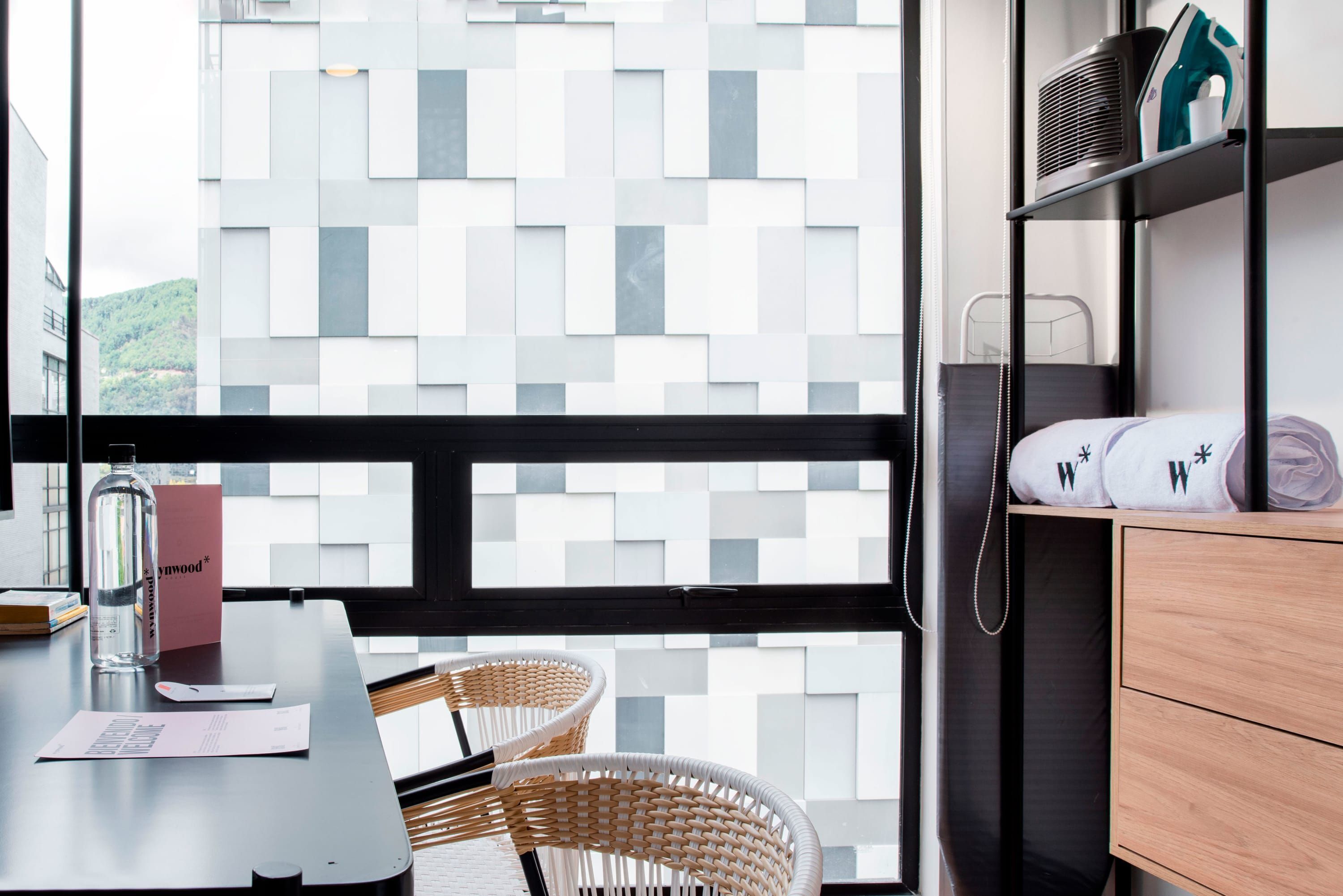 A modern hotel room with a large window, a wooden dresser, a wicker chair, and a table with a bottle and papers.