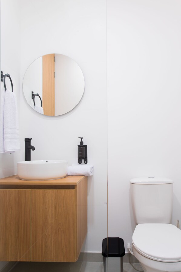 Une salle de bain moderne avec un meuble-lavabo en bois, un miroir rond, un robinet noir, des toilettes blanches et une poubelle noire.