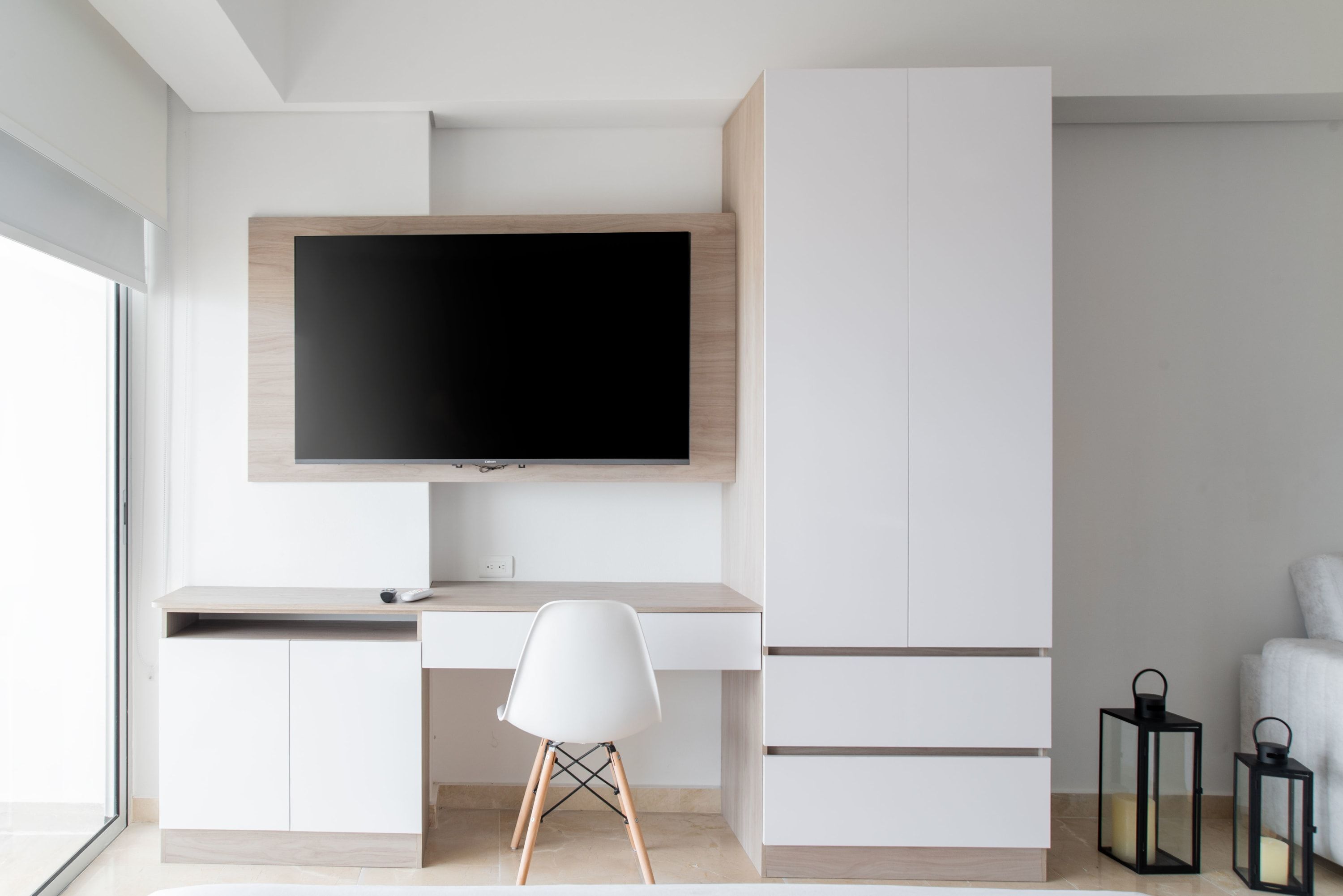 Quarto moderno e minimalista com uma televisão de tela plana grande na parede, uma escrivaninha branca com cadeira e um guarda-roupa embutido.