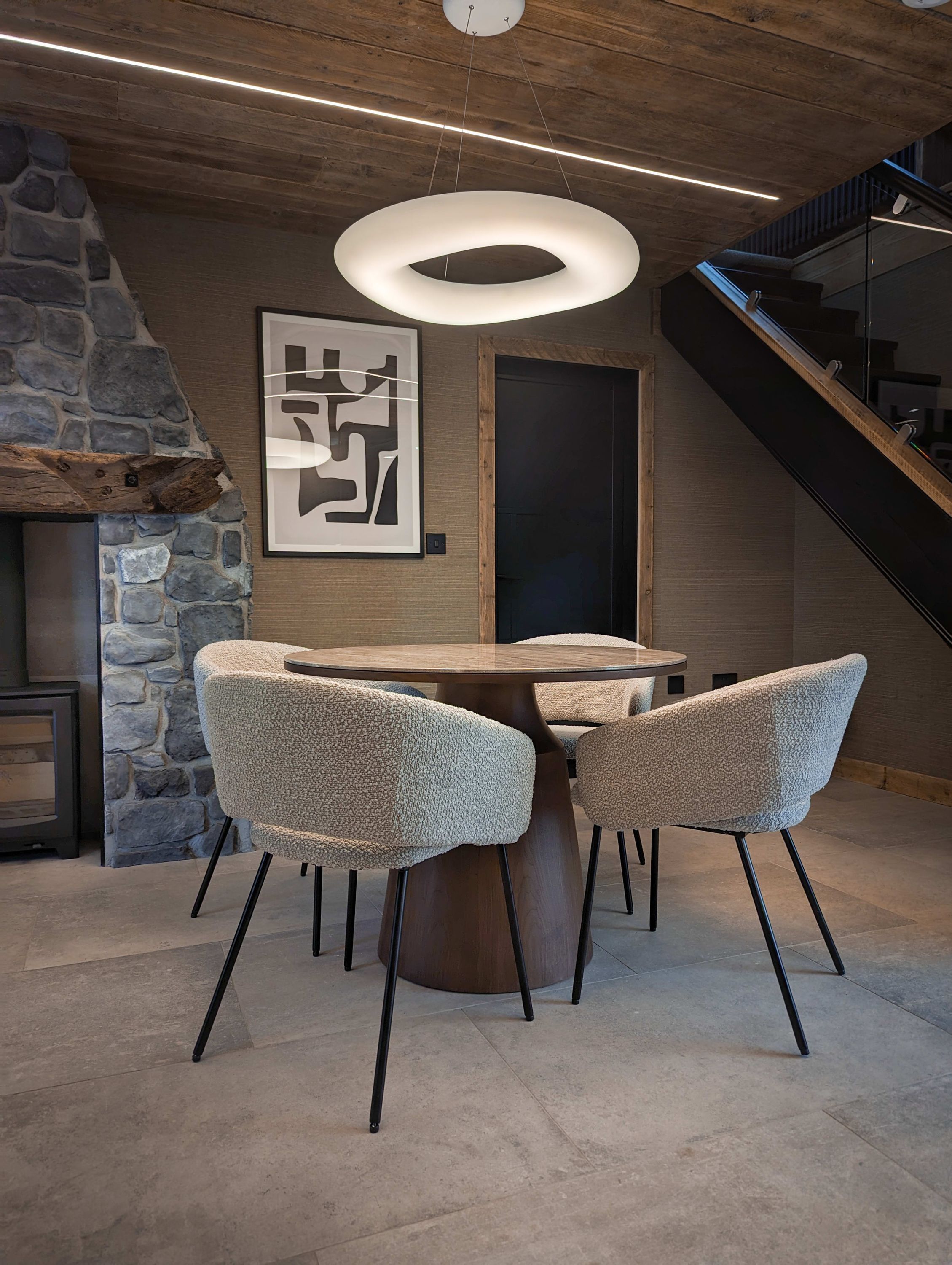 A modern dining area with a round table and four chairs, a stone wall, a wooden ceiling, and a staircase.