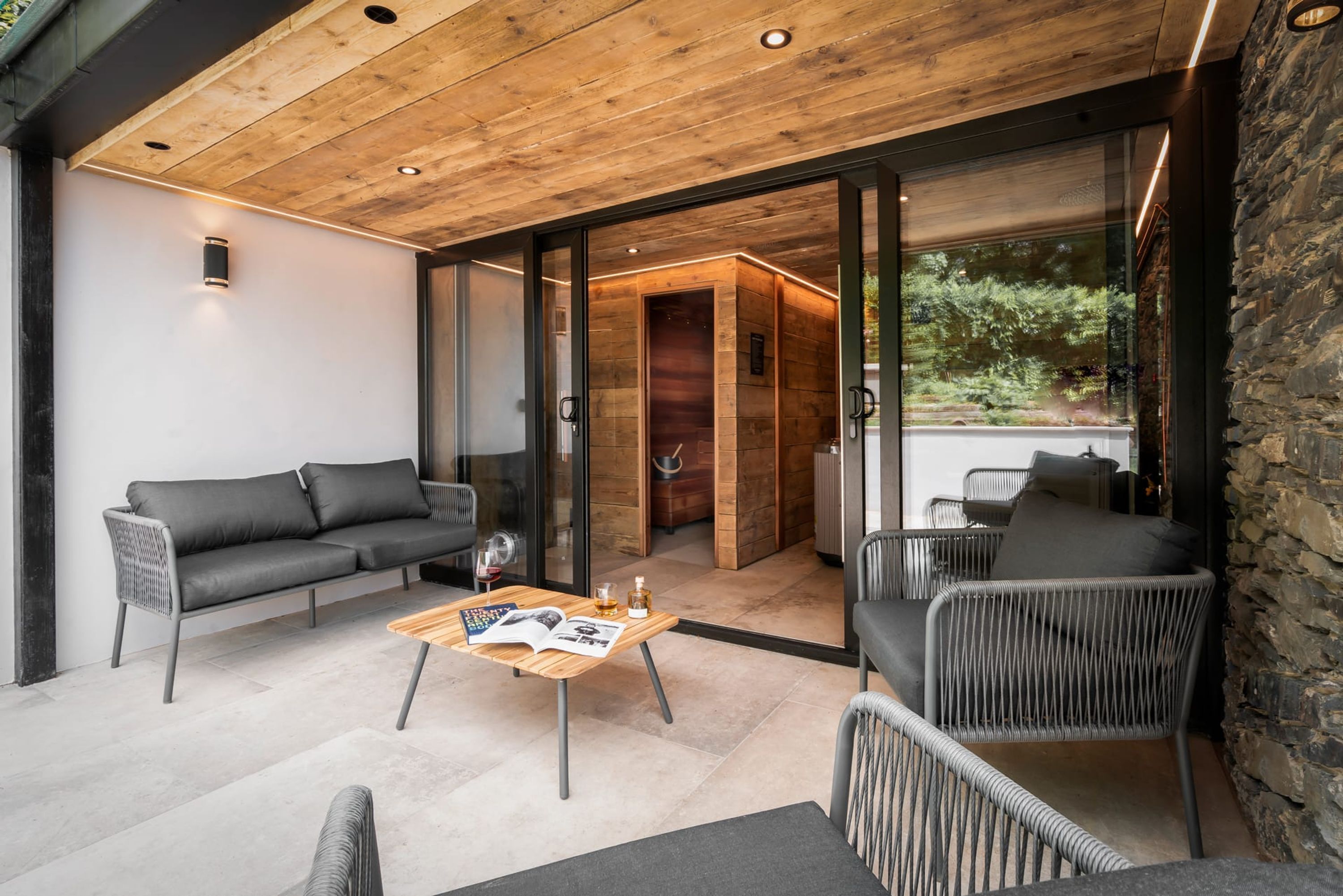 A modern outdoor seating area with a wooden ceiling, grey upholstered sofas, and wicker chairs.
