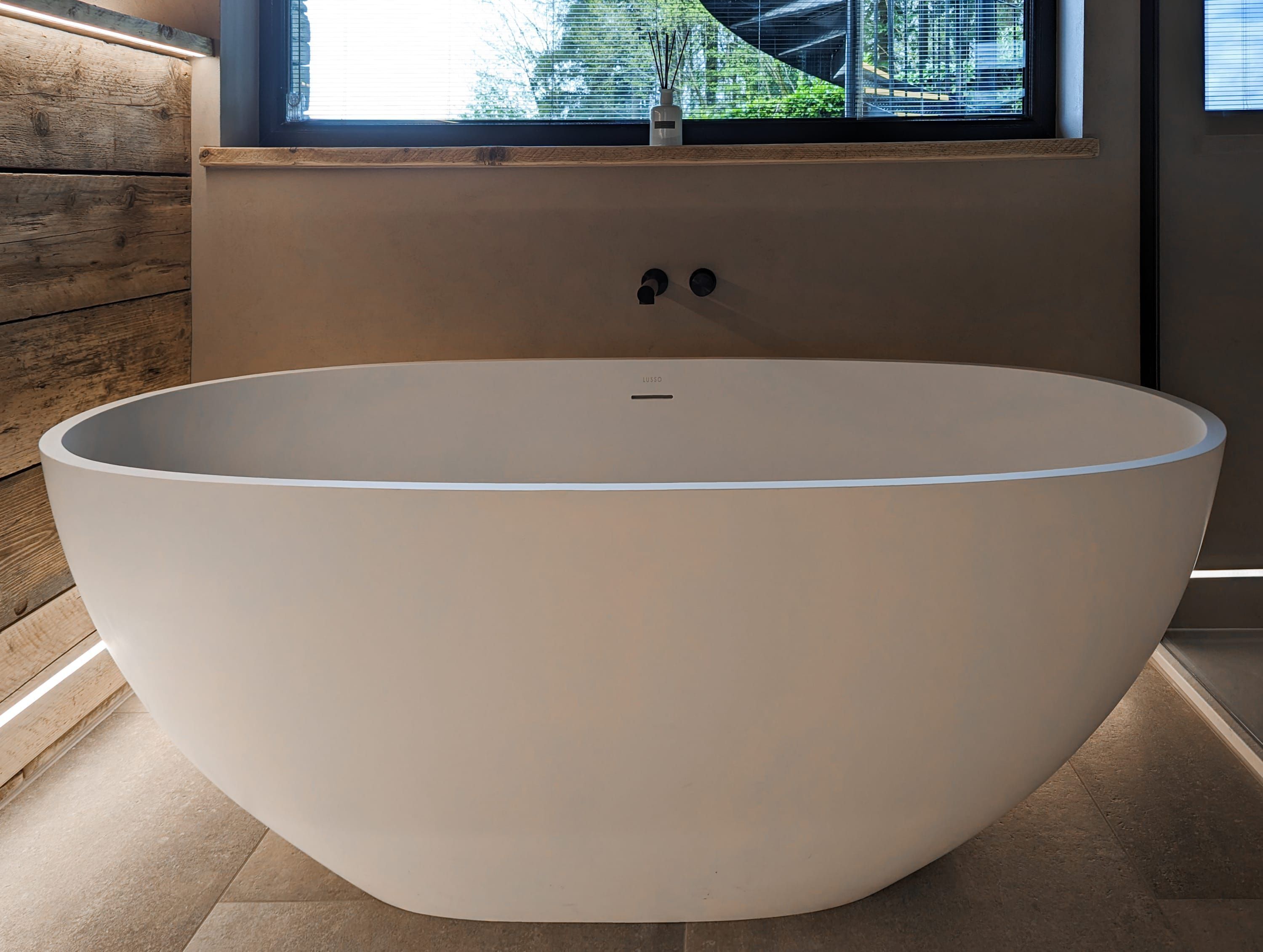 A large, white, freestanding bathtub in a modern bathroom with a wooden wall and a window above it.