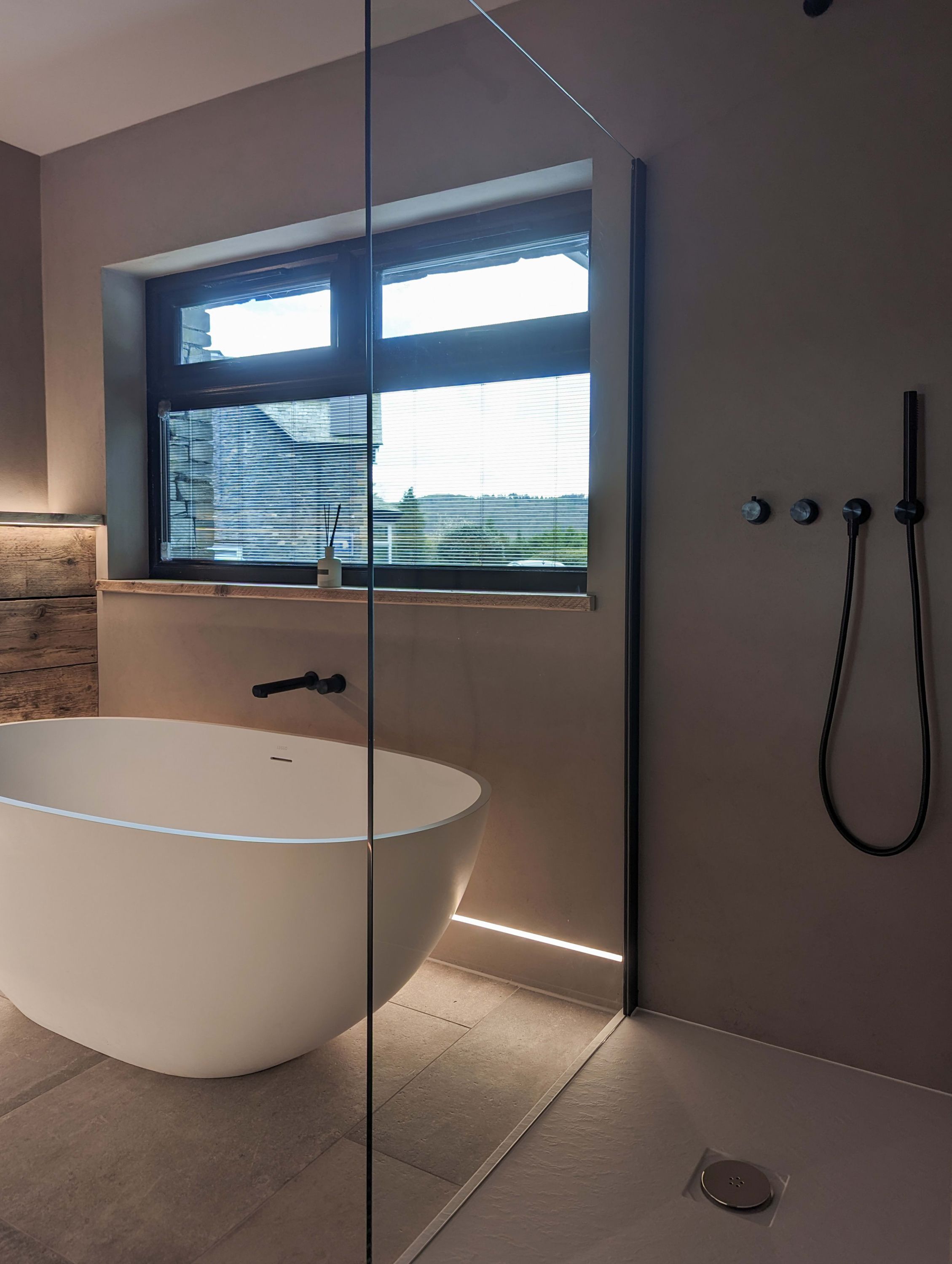 A modern bathroom with a freestanding bathtub, a window with blinds, and a shower area.