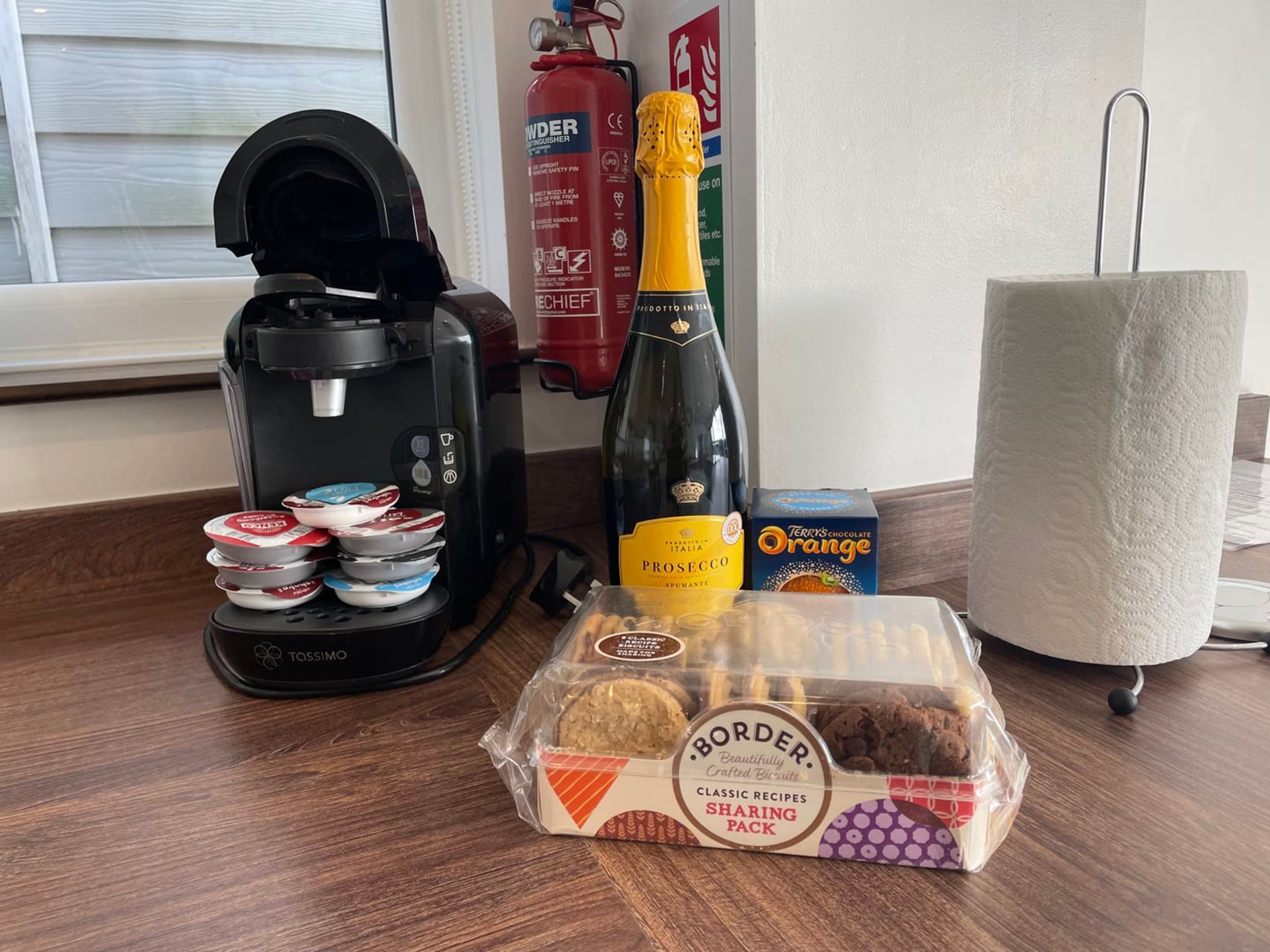 A coffee maker, a bottle of Prosecco, a box of Terry's Orange chocolate, a paper towel roll, and a Border biscuits sharing pack on a wooden table.