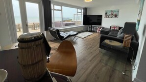 A spacious living room with a large window overlooking the beach, a flat-screen TV, and a cozy seating area with a sofa and armchairs. - Exquisite Beachfront Residence With Luxurious Interiors The Cozy Clam Pevensey Bay (Pevensey)