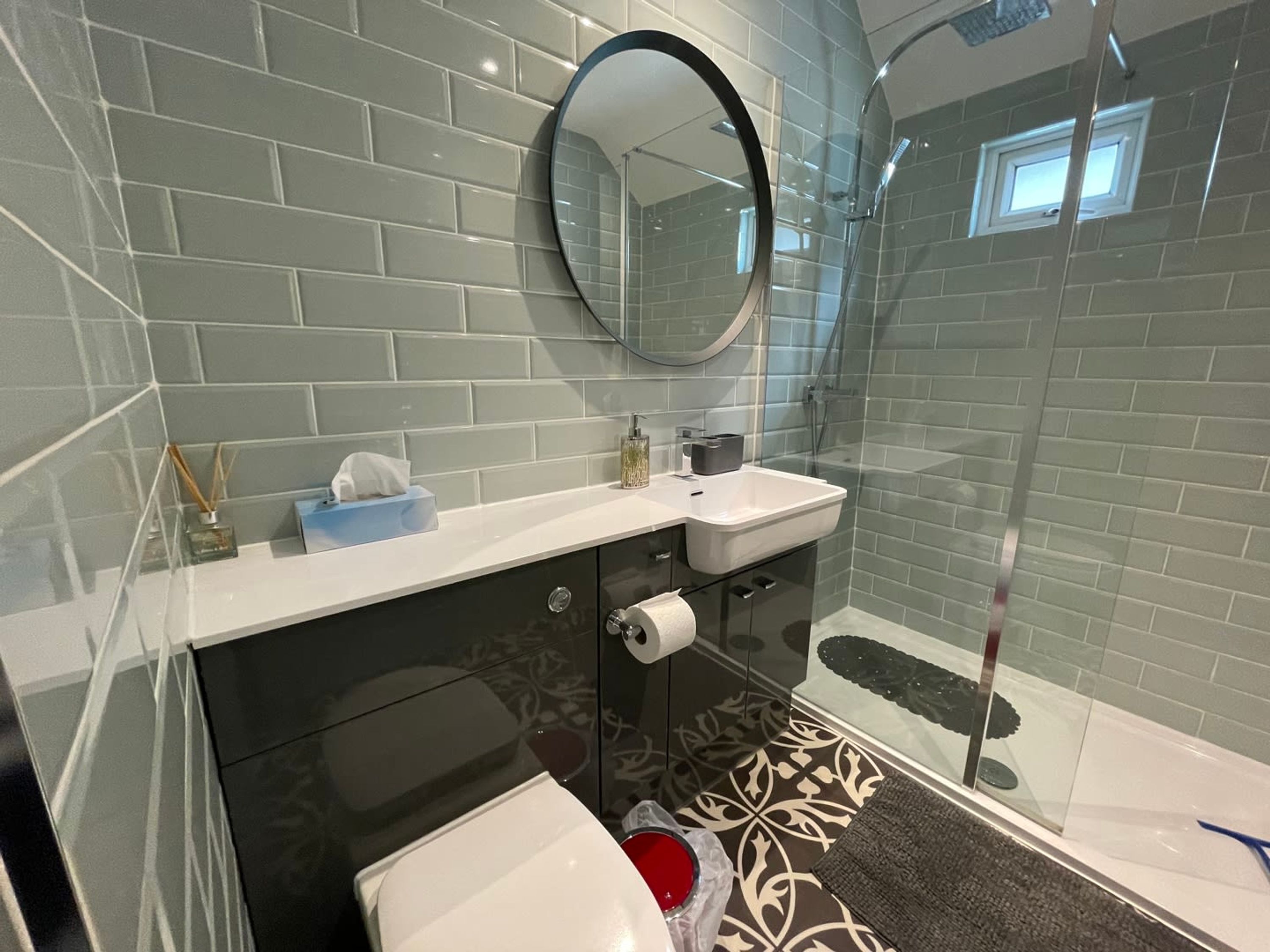 A modern bathroom with grey tiled walls, a round mirror, a white sink, a black cabinet, and a glass shower enclosure.