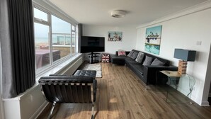 A modern living room with a large window, a flat-screen TV, a black leather sofa, a small table, and a lamp. - Exquisite Beachfront Residence With Luxurious Interiors The Cozy Clam Pevensey Bay (Pevensey)