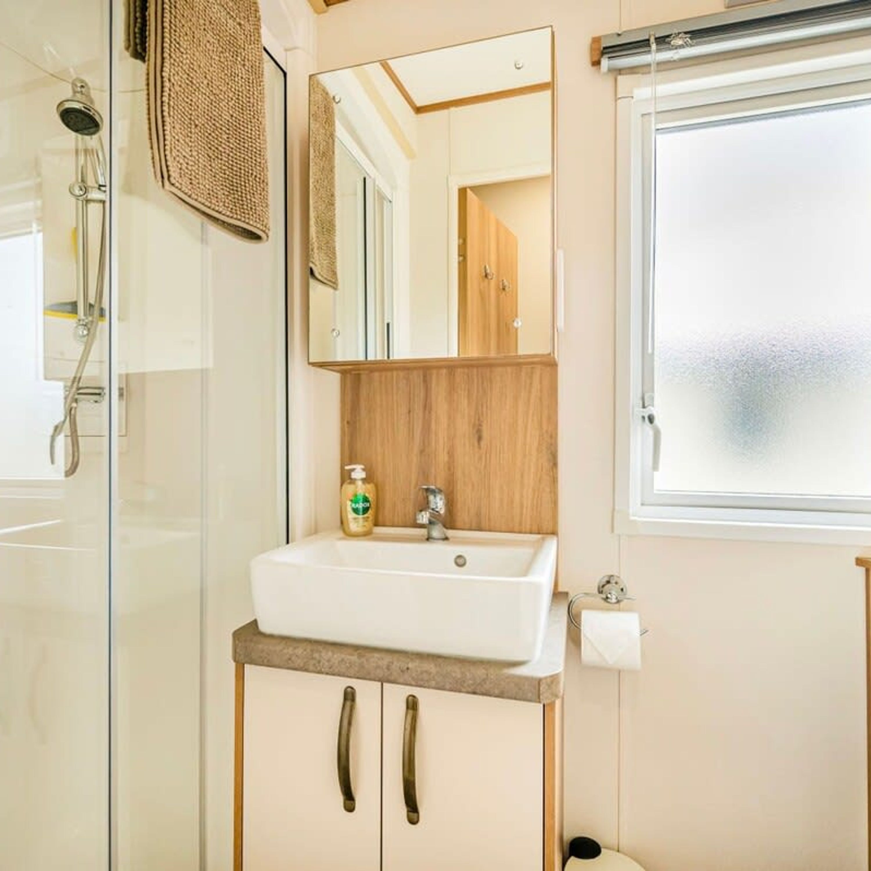 A bathroom with a glass shower enclosure, a white sink with a wooden cabinet, a mirror, and a window.