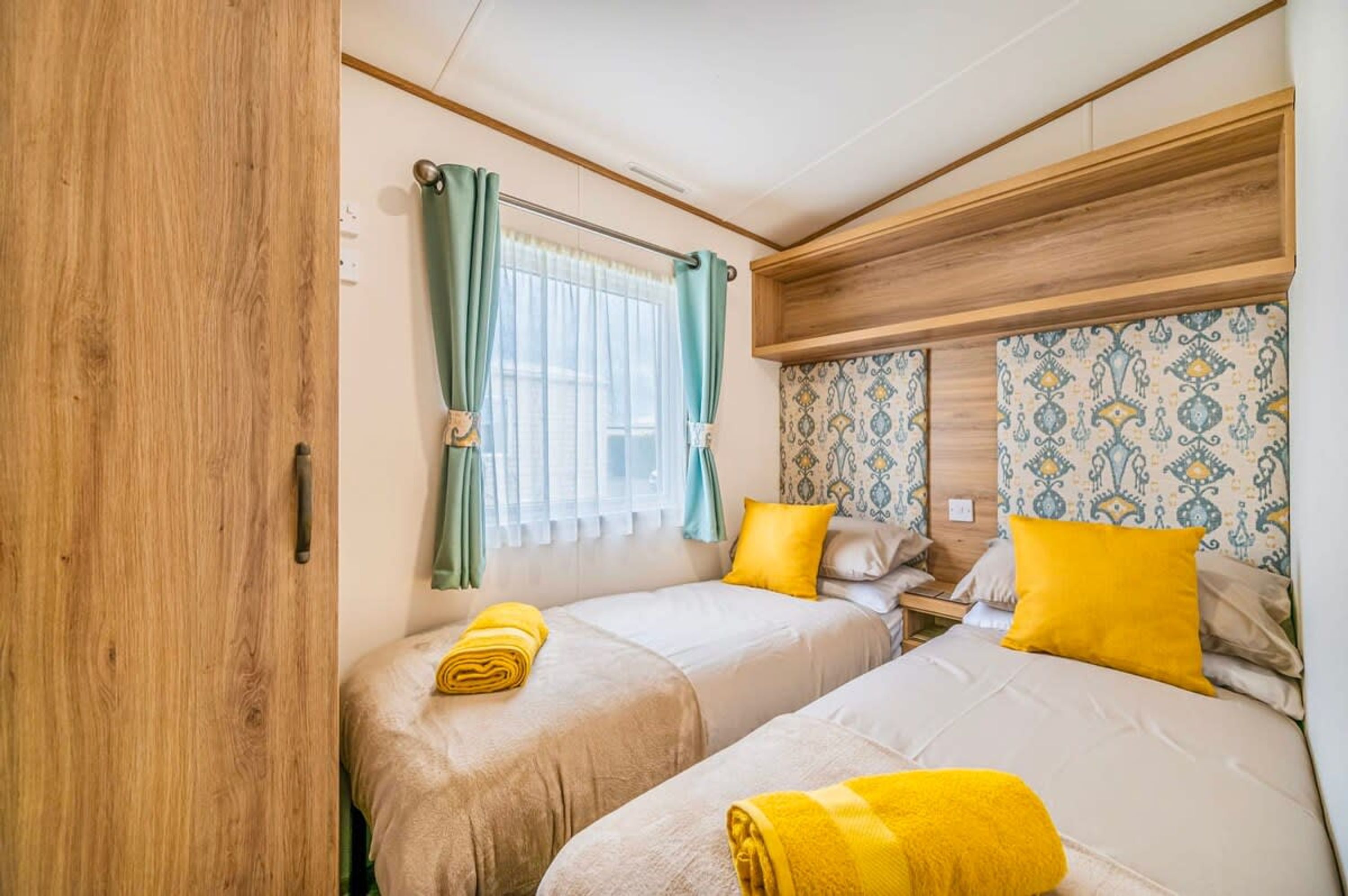 A room with two beds, a wooden wardrobe, a window with curtains, and patterned wallpaper.