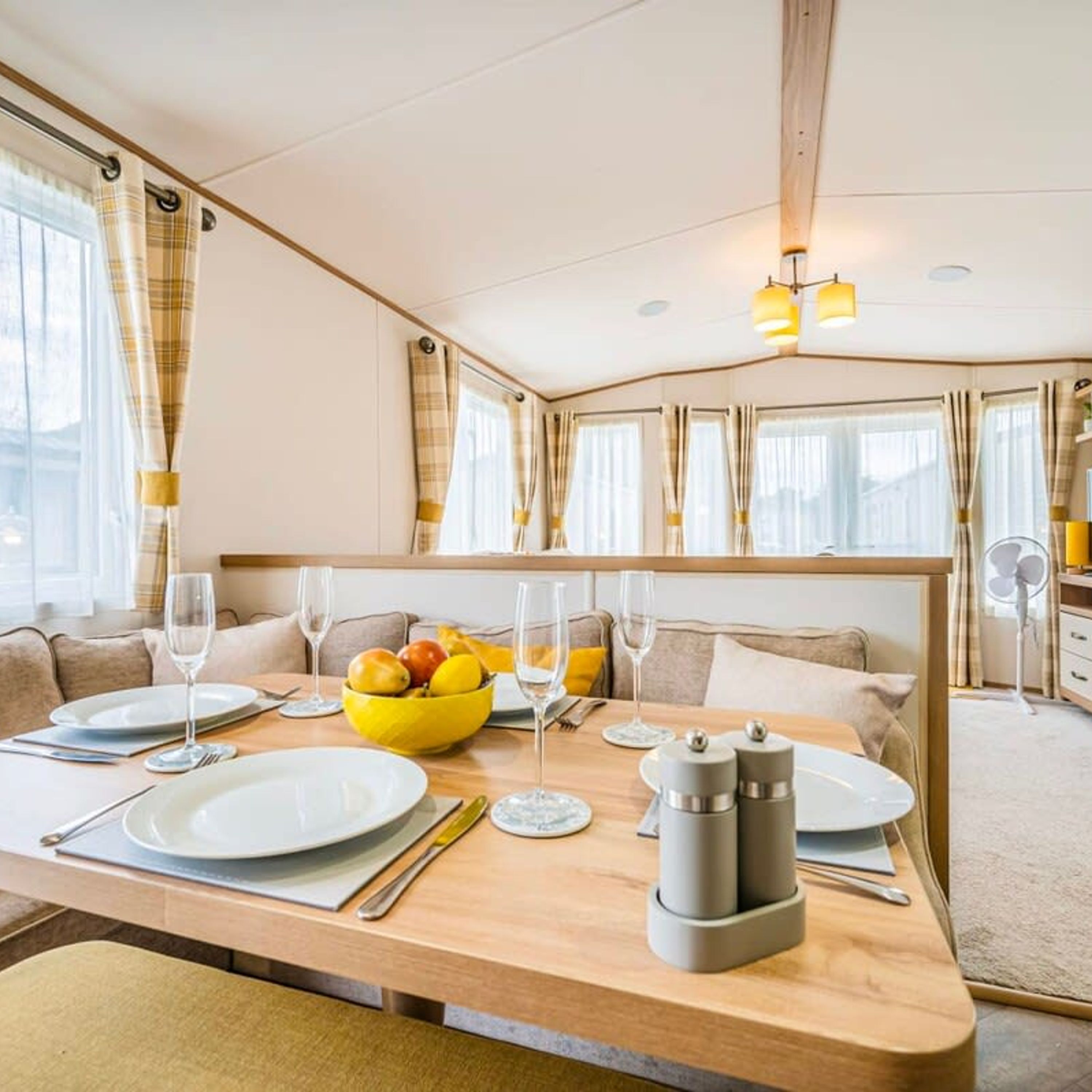 Un coin repas avec une table en bois dressée pour deux personnes, un saladier de fruits et un ventilateur en arrière-plan.