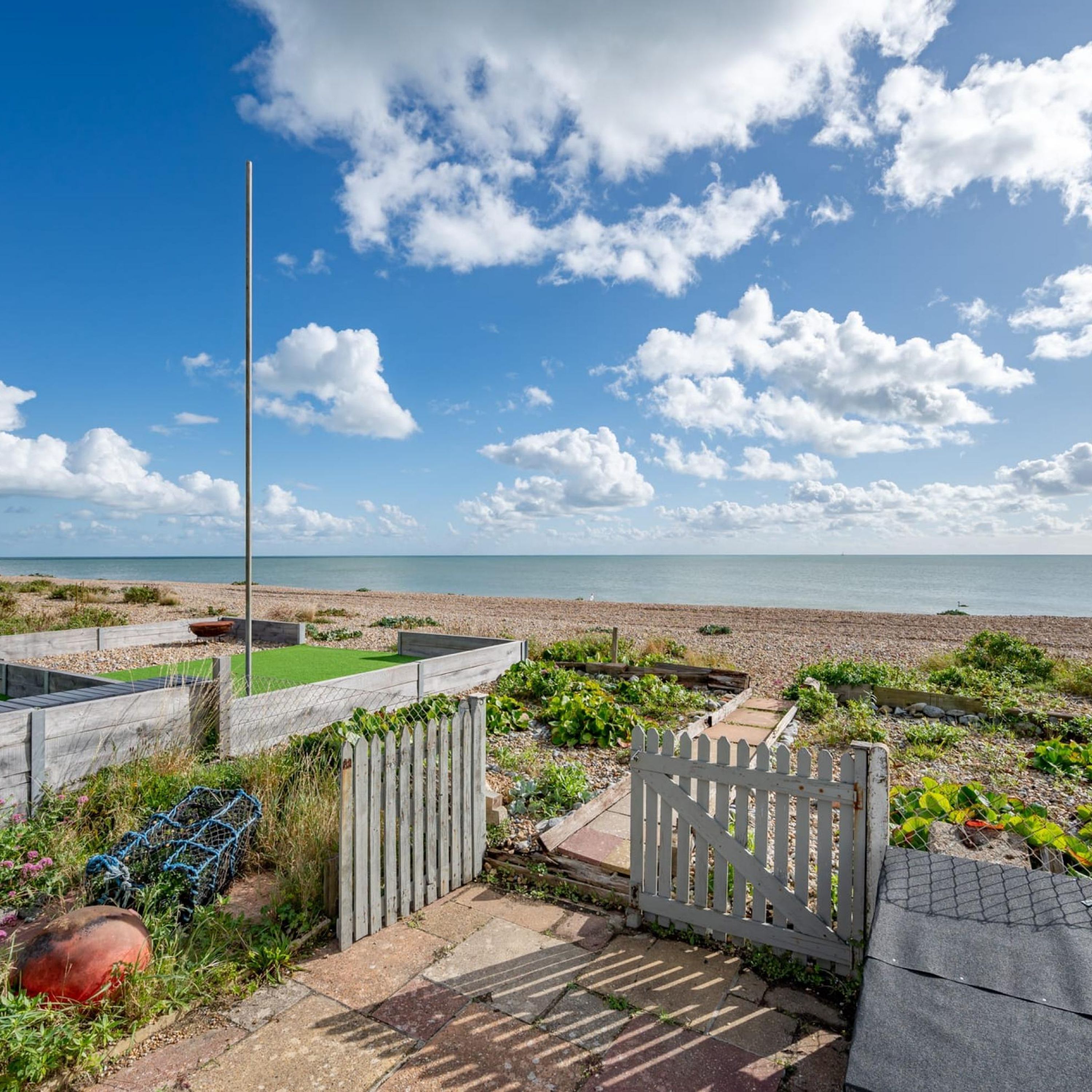 A coastal garden with a wooden gate, a flagpole, and a pebble beach in the background.