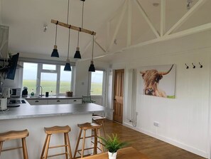 A spacious kitchen with a central island, wooden stools, pendant lighting, and a large window offering a view of the outdoors. - Cosy Beach Cottage Metres From The Beach Jurys Gap Camber Sands (Rye)