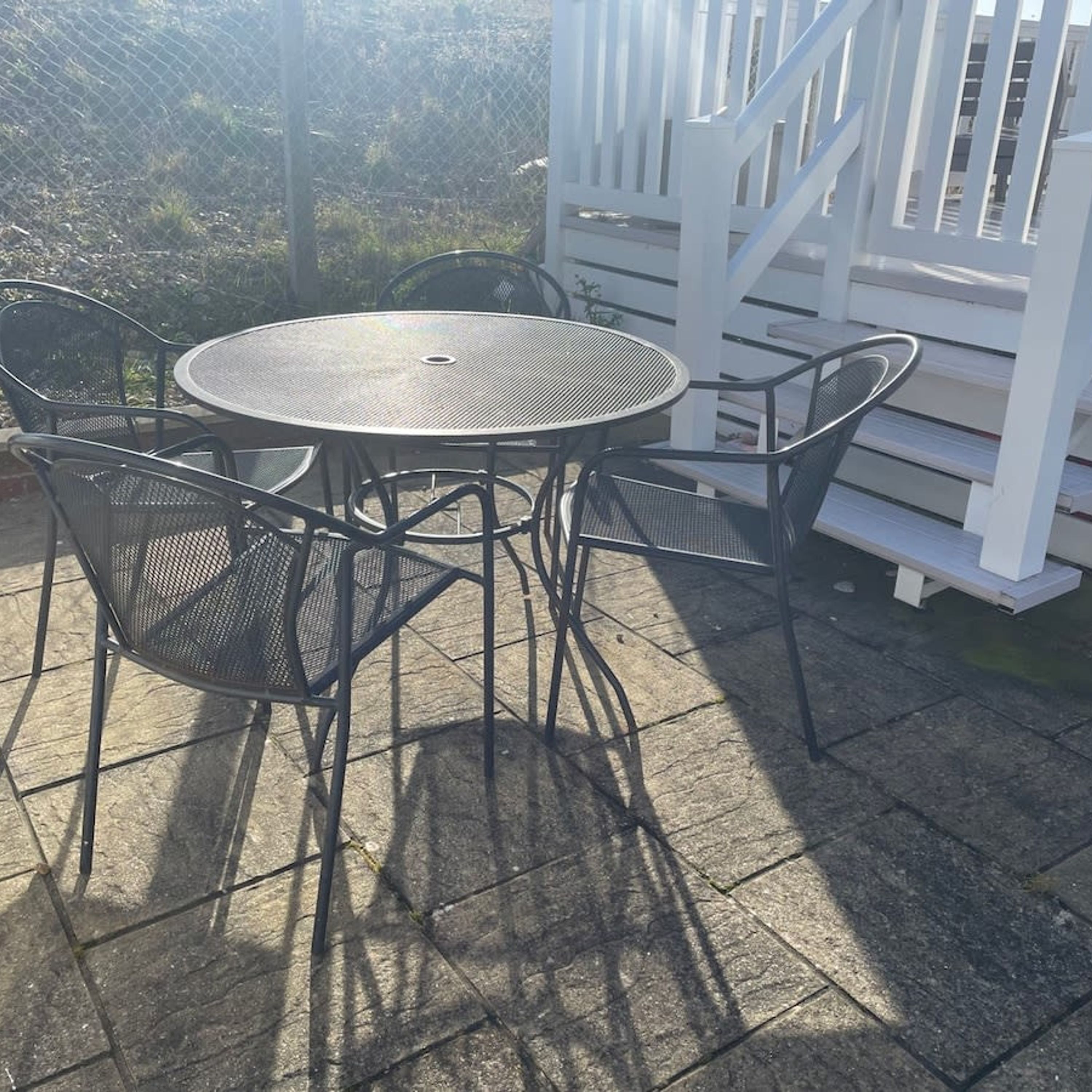 Une table ronde en métal, entourée de quatre chaises, est placée sur une terrasse en pierre, avec une clôture blanche en arrière-plan.