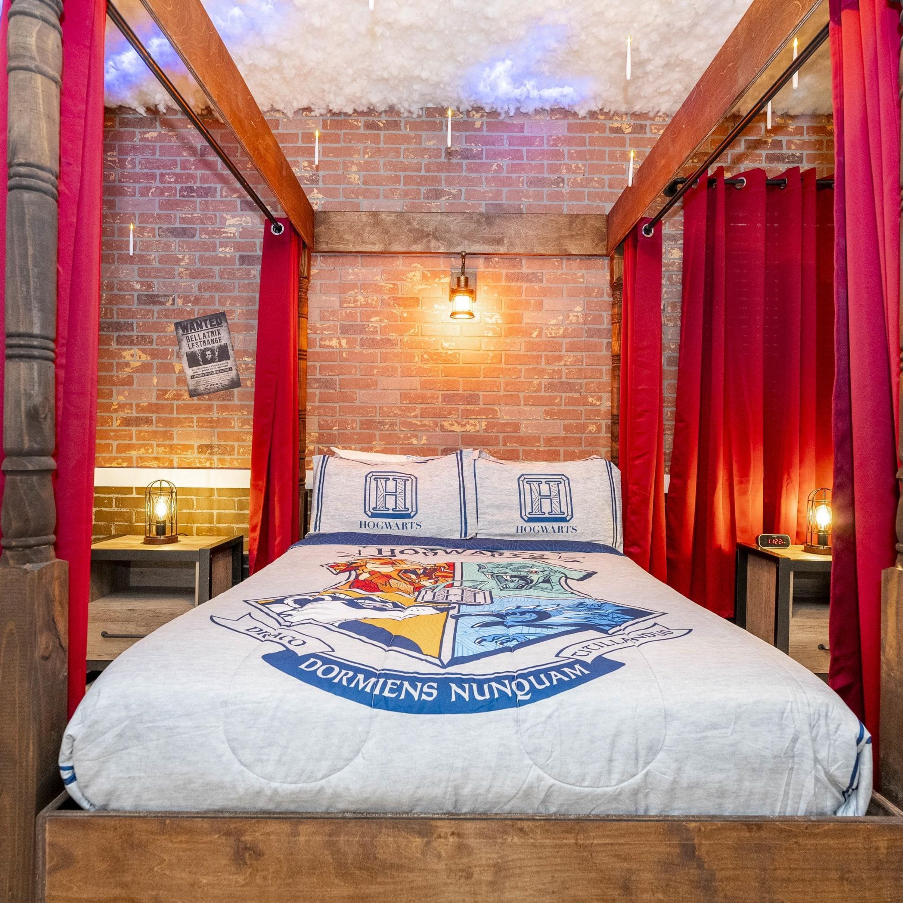 A room with a brick wall, a bed with a Hogwarts-themed duvet, red curtains, and a small table with a lamp.
