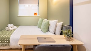 Un dormitorio ordenado con una cama, un escritorio con una computadora portátil, una ventana con un reloj y una lámpara de pared.