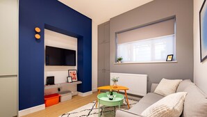 Una sala moderna con una pared azul destacada, un televisor de pantalla plana, un sofá, una mesita con una botella y una ventana con persianas.