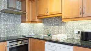 A compact kitchen with wooden cabinets, a stainless steel oven, a dishwasher, and a tiled backsplash. - Ferncourt Stepaside (Dublin)