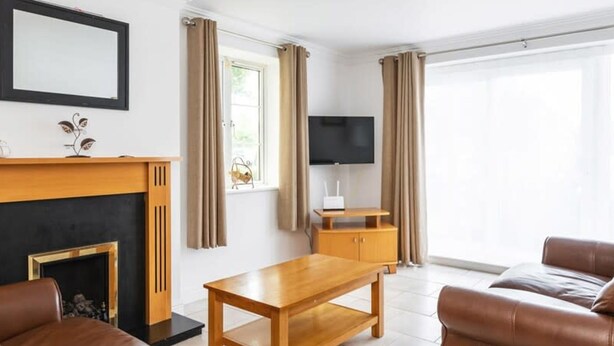 A living room with a fireplace, leather sofas, a wooden coffee table, a TV, and a large window with curtains. - Ferncourt Stepaside (Dublin)