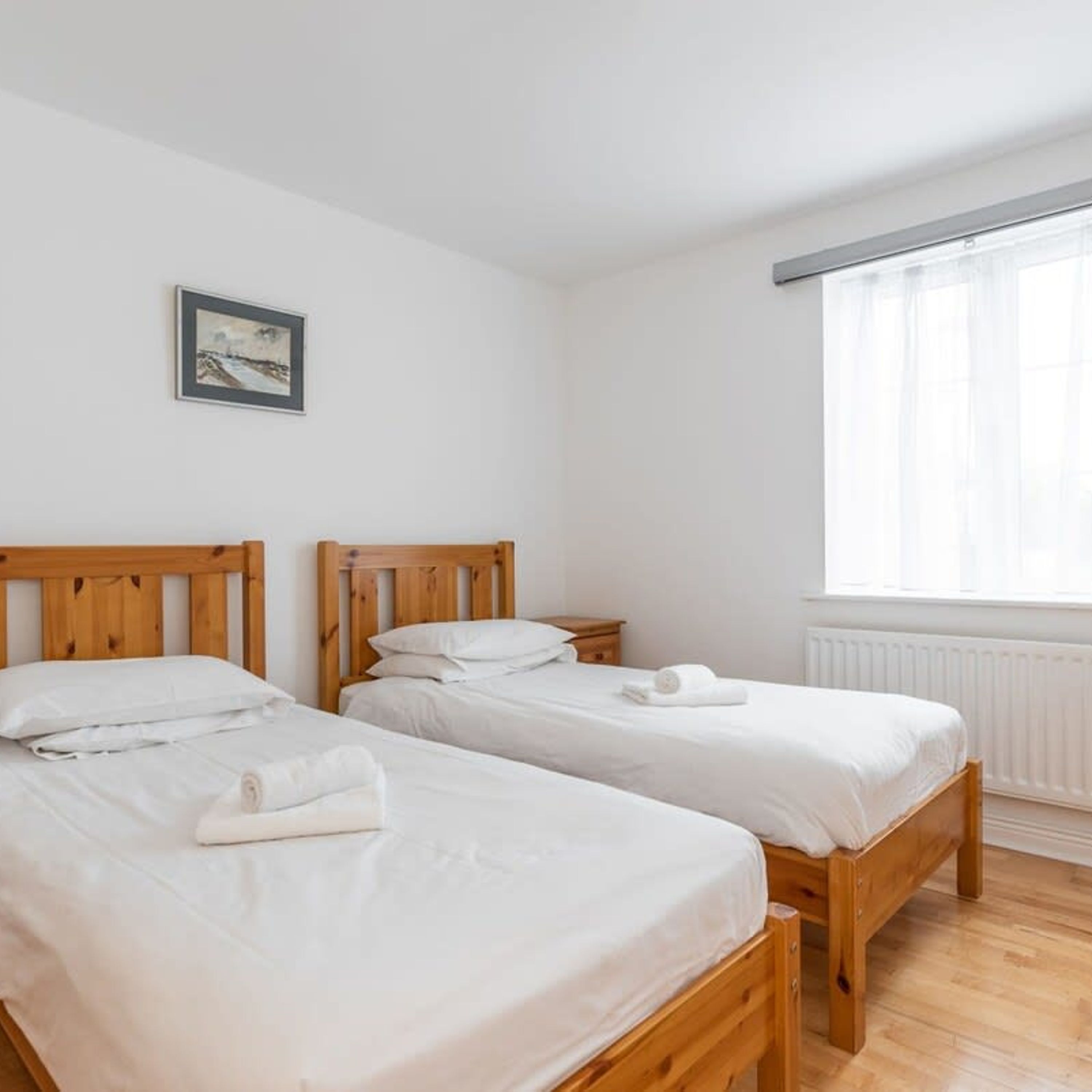 A room with two wooden beds, white bedding, a framed picture on the wall, and a window with sheer curtains.