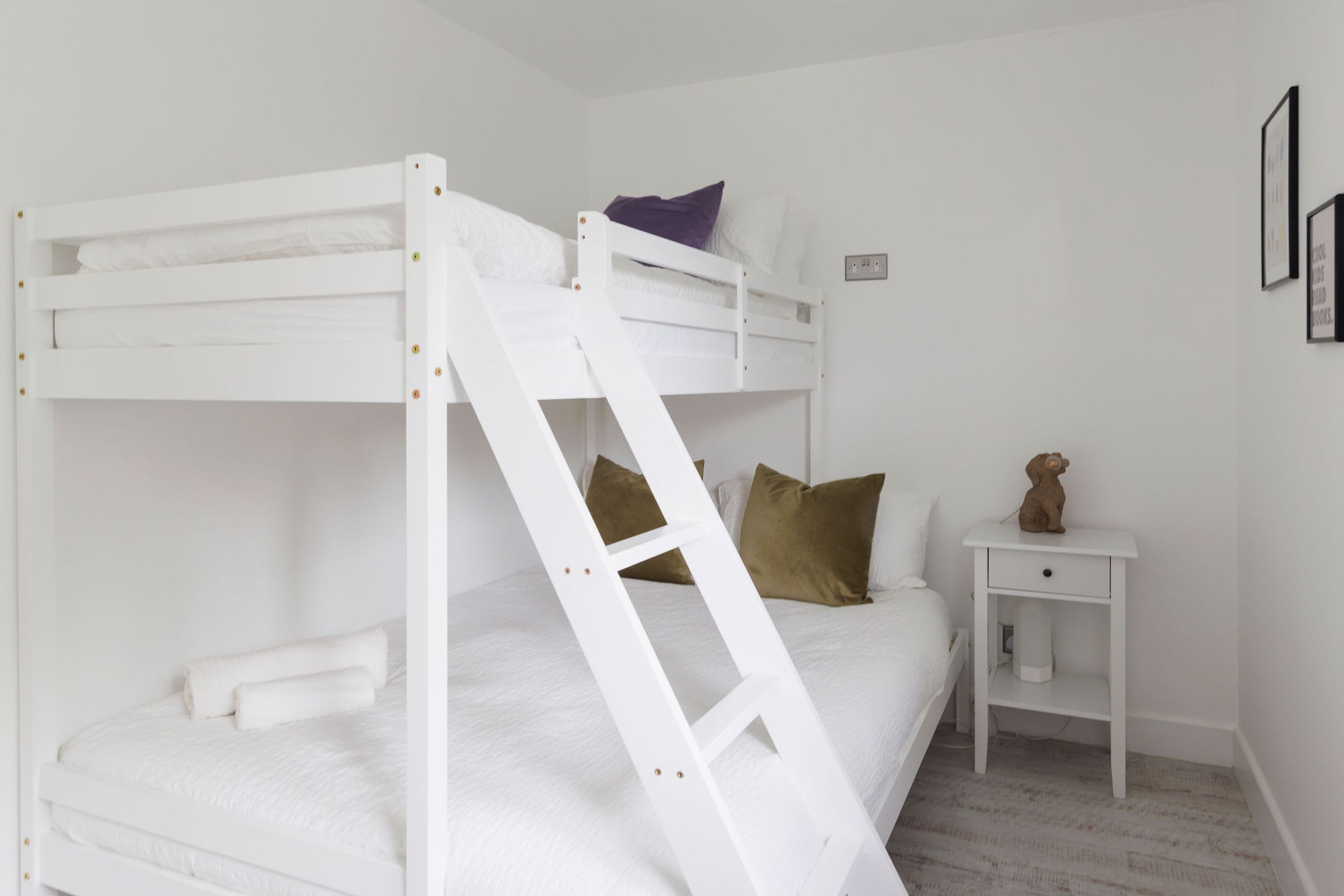 Una cama litera con escalera, ropa de cama blanca, una mesita de noche pequeña con un oso de peluche y un interruptor de luz en la pared.