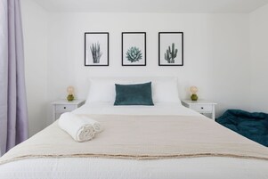 Una cama bien hecha con edredón blanco y una toalla blanca enrollada, flanqueada por dos mesitas de noche con lámparas, y adornada con estampados botánicos en la pared.