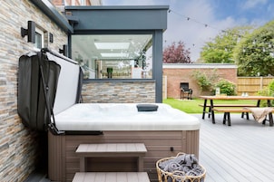 Una bañera de hidromasaje con una terraza de madera, una cesta de mimbre con toallas y una mesa y banco de madera al fondo.