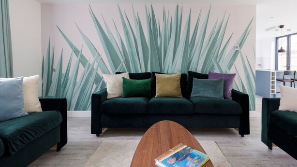 Un soggiorno moderno con un divano verde scuro, un tavolino in legno e un grande murale con foglie verdi.