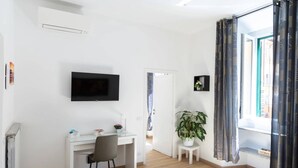 Une pièce moderne et bien éclairée, avec un bureau blanc, une chaise et un téléviseur à écran plat fixé au mur. Une plante en pot est posée sur une petite table à côté du bureau, et une fenêtre est agrémentée de rideaux à motifs.