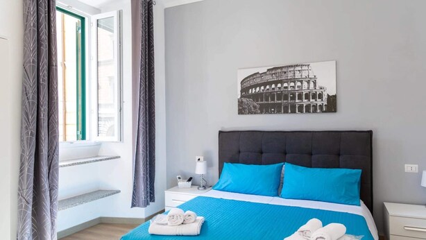 Une chambre d’hôtel avec un lit, une table de chevet, une fenêtre avec des rideaux et un tableau représentant le Colisée accroché au mur.