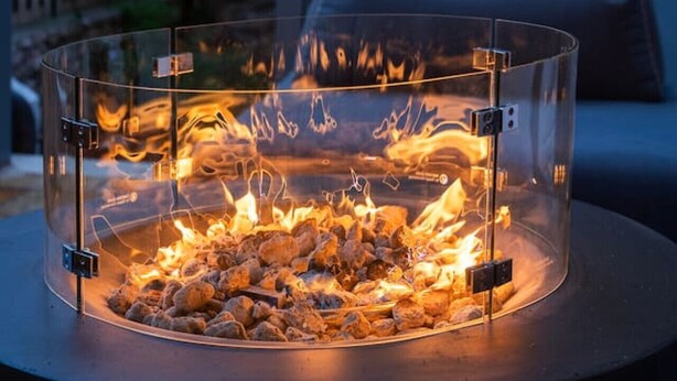 Ein in Glas eingefasster Feuerkorb mit Metallrahmen, umgeben von einem dunklen, runden Tisch.