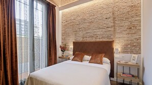A hotel room with a large bed, a brick wall, a nightstand with a lamp, and a small table with a book. - Pau Claris Double Home Next to Paseo de Gracia (Barcelona)