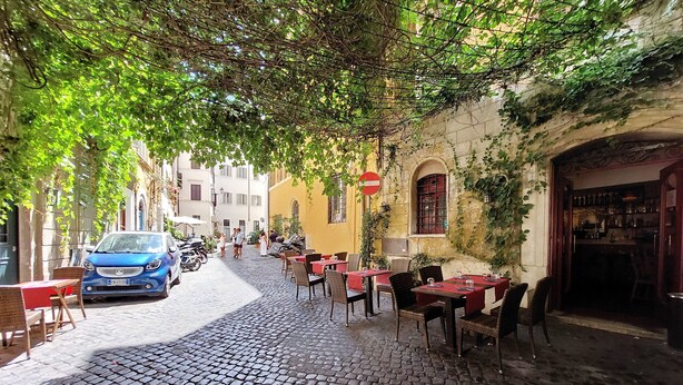 Une rue pavée avec des tables et des chaises installées à l’extérieur, et une voiture bleue garée sur le côté gauche.