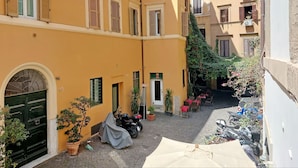 Une cour étroite avec un bâtiment jaune, une porte noire, des plantes en pot et des motos garées.