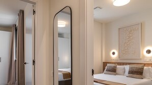 Camera d'albergo con un grande specchio, un letto con biancheria da letto bianca e dettagli marroni, una testiera con luci a parete e un'opera d'arte incorniciata sopra il letto.