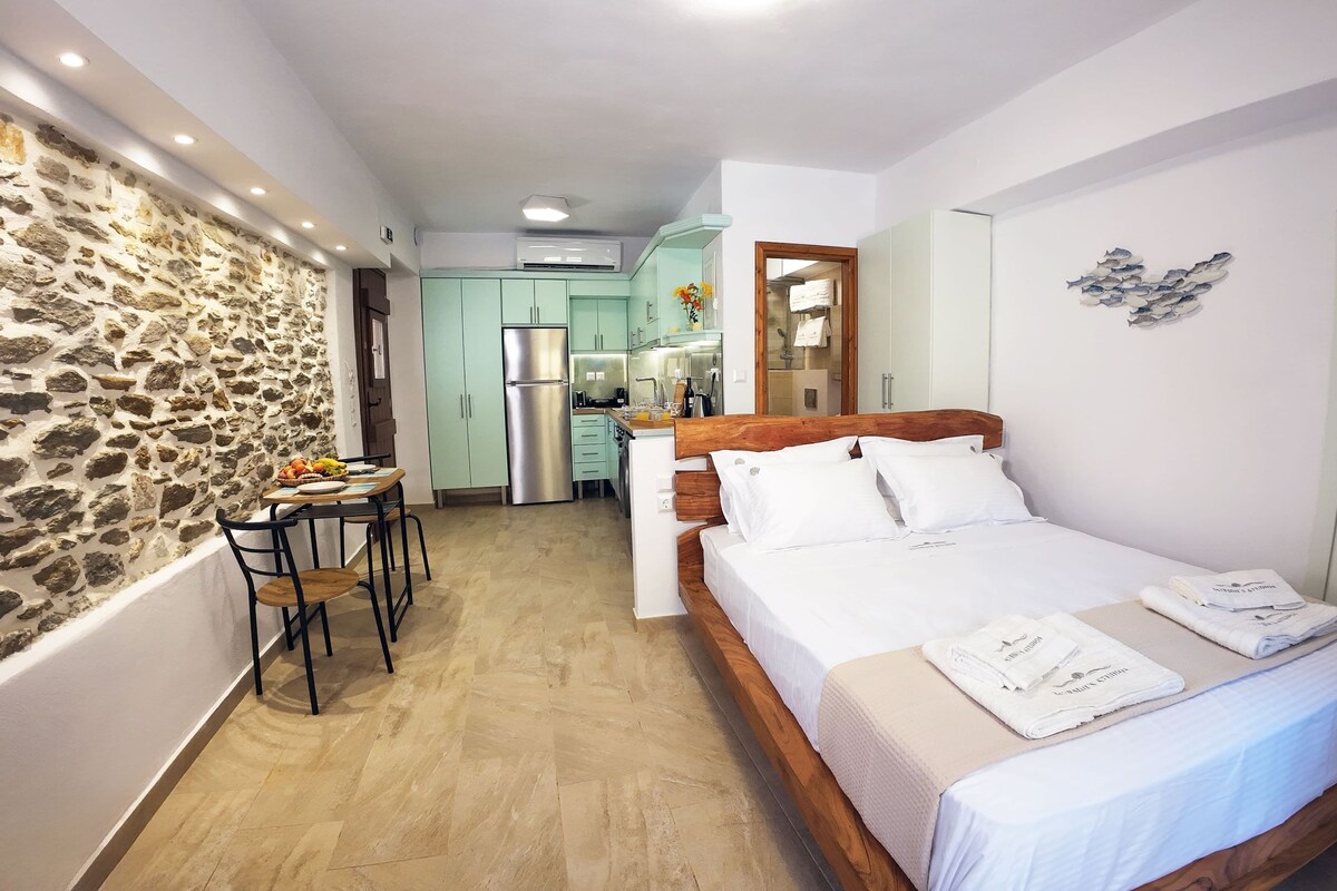 Une chambre comprenant un lit, une tête de lit en bois, une table de chevet avec des serviettes, un mur en pierre, un coin cuisine et une salle de bain avec des toilettes et un lavabo.