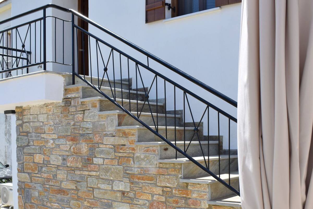A stone staircase leading to a white building with wooden shutters, a wooden table with a cushion and a lantern, and a chair with a leaf pattern.