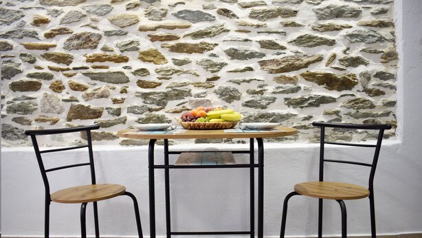 Una habitación con pared de piedra, una mesa redonda de madera, dos sillas de metal y una cesta de frutas sobre la mesa.